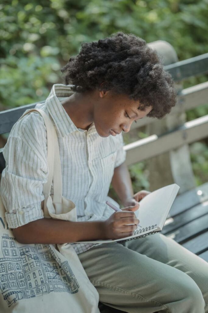 Woman sittted In a park bench doing self-discovery journaling prompts.