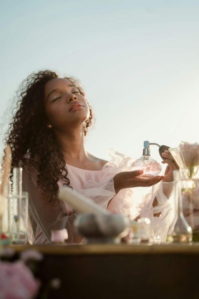 Image of a woman who's wearing a pink dress and spraying perfume on herself, sitting in an open field, signifying a feminine energy microhabit.