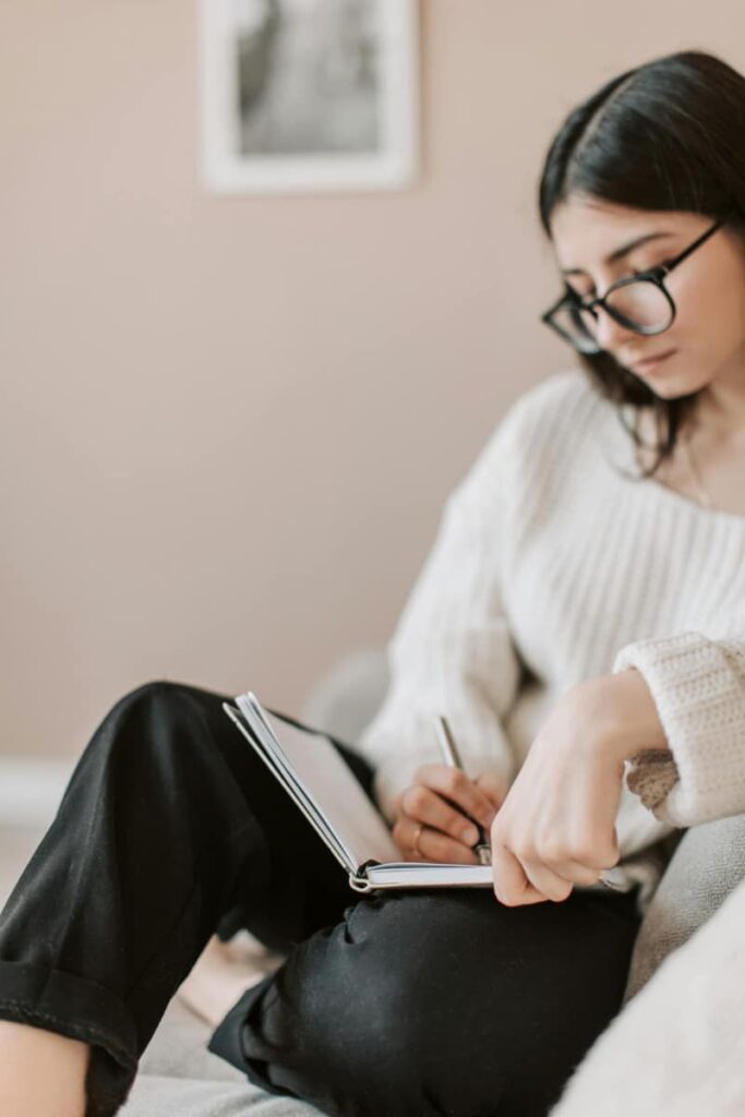 Image of a woman in a white sweater and sunglasses is seated and writing in a black journal using journaling prompts for letting go.