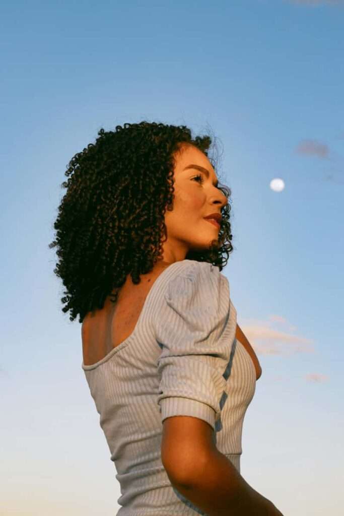 Image of a woman with curly hair and a grey top standing outside looking very satisfied representing the benefits of using the correct manifestation methods for your personality.