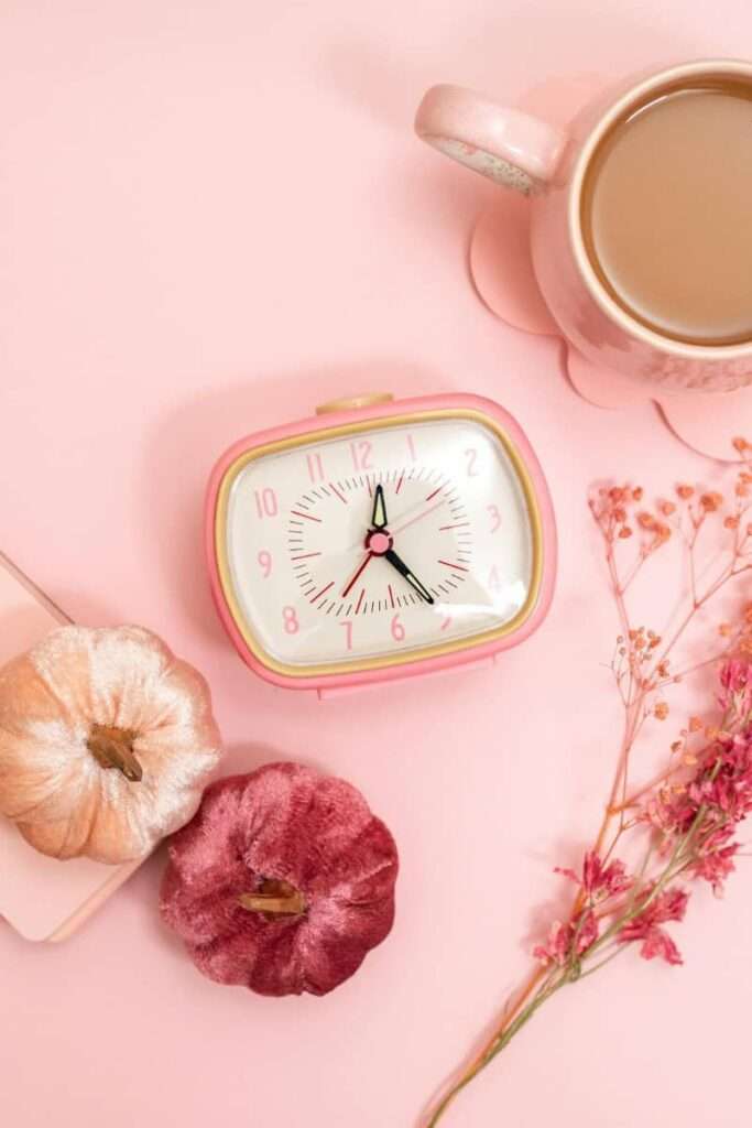 A pink clock on a table with flowers to indicate time management as a self-improvement tool for women 
