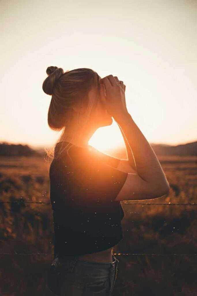 Image of a woman hiding her face, standing before a sunset, casting a shadow on her, representing shadow work.