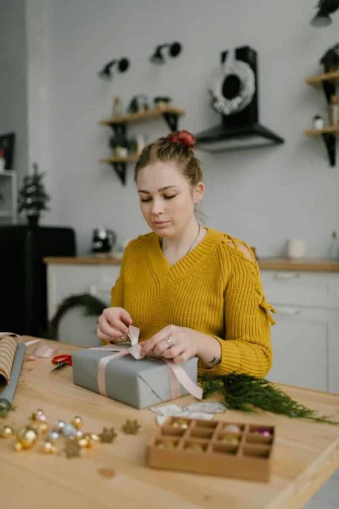 Image of a woman in a yellow sweater seated at a table wrapping presents representing the festivities.