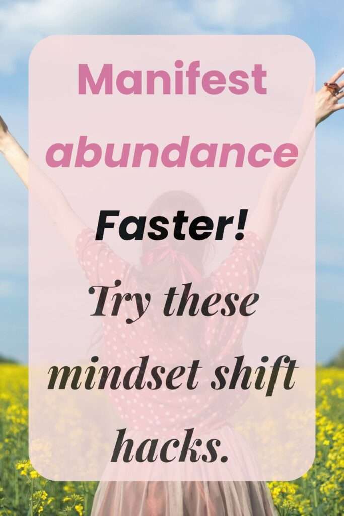 Pink Pinterest pin with a Jovial woman in the background in a flower field with her hands up and an overlay text reading "manifest abundance faster. Try these mindset shift hacks.